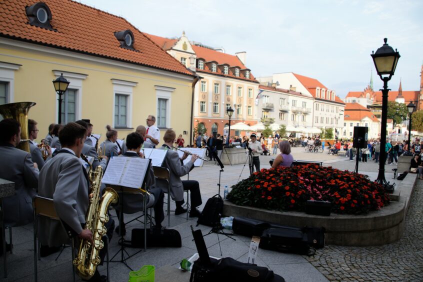 Orkiestra, dyrygent, prowadząca, w oddali uczestnicy koncertu na tle kamienic i katedry