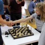 Uścisk dłoni uczestników partii szachowej, w tle uczeń-sędzia, kibic i inni uczestnicy turnieju