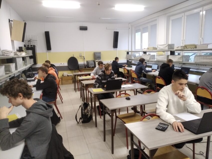 Uczniowie siedzą przy stolikach z laptopami w sali lekcyjnej .