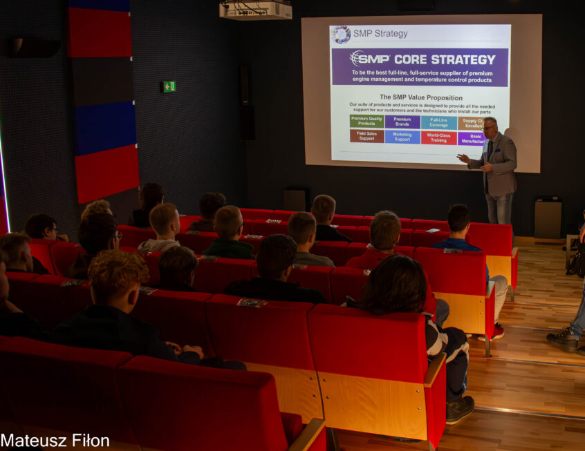 Przedstawiciel firmy SMP Poland stoi przy wyświetlonym slajdzie z prezentacji, na fotelach siedzą uczniowie