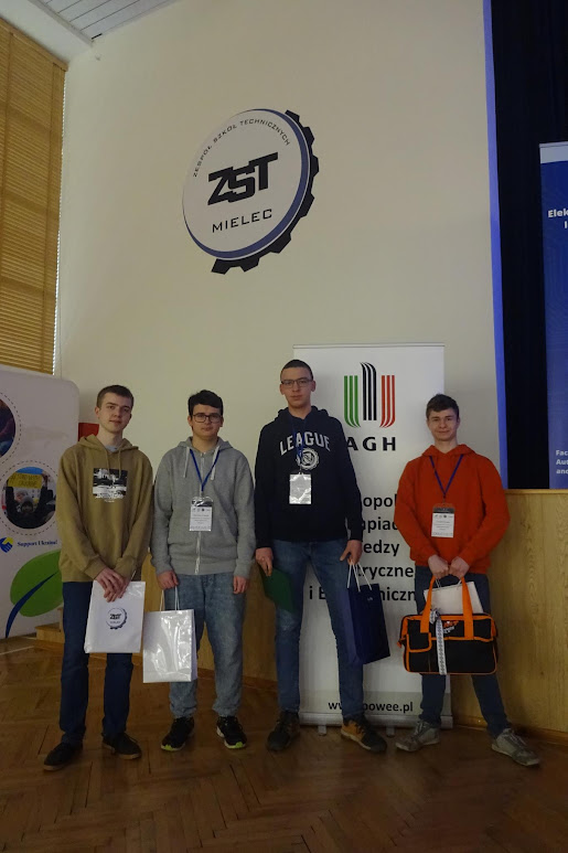 4 uczniów z prezentami stoi na tle rollupu AGH i logo ZST Mielec